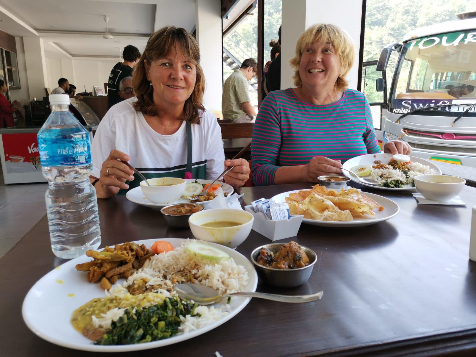 Two women enjoying traditional meals at a restaurant table, smiling with food and drinks around.